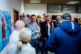 Gruppe von Menschen in einem Raum, einige stehen mit verschränkten Armen, eine Person trägt eine blaue Jacke und eine Kappe