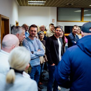Gruppe von Menschen in einem Raum, einige stehen mit verschränkten Armen, eine Person trägt eine blaue Jacke und eine Kappe