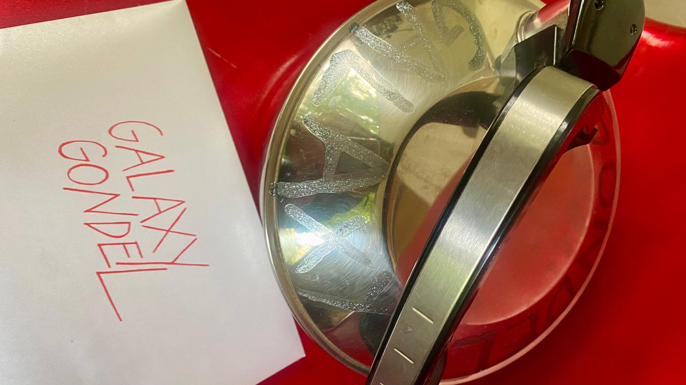 Ältere, metallische Teekanne mit Griff auf rotem Untergrund neben einem weißen Zettel mit handschriftlicher roter Aufschrift 'GALAXY GONDEL'