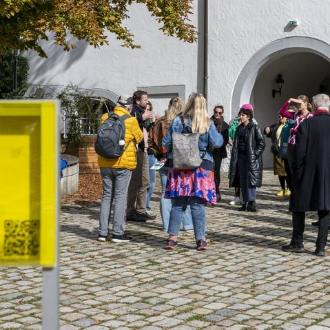 Die Gruppe der Teilnehmer*innen im Gespräch mit den Festival-Kurator*innen Herbordt/Mohren. Die Gruppe steht bei strahlendem Sonnenschein im Hof von Schloss Waldenbuch. Sie Sonne scheint. Herbstlich verfärbte Blätter eines Baumes ragen von oben in das Bild. Im Vordergrund steht ein gelbes, transparentes Schild mit einem QR-Code.
