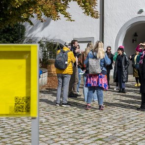 Die Gruppe der Teilnehmer*innen im Gespräch mit den Festival-Kurator*innen Herbordt/Mohren. Die Gruppe steht bei strahlendem Sonnenschein im Hof von Schloss Waldenbuch. Sie Sonne scheint. Herbstlich verfärbte Blätter eines Baumes ragen von oben in das Bild. Im Vordergrund steht ein gelbes, transparentes Schild mit einem QR-Code.