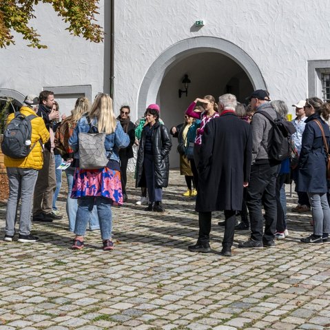 Die Gruppe der Teilnehmer*innen im Gespräch mit den Festival-Kurator*innen Herbordt/Mohren. Die Gruppe steht bei strahlendem Sonnenschein im Hof von Schloss Waldenbuch. Sie Sonne scheint. Herbstlich verfärbte Blätter eines Baumes ragen von oben in das Bild. Im Vordergrund steht ein gelbes, transparentes Schild mit einem QR-Code.