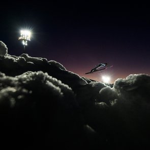 Skispringer in der Luft vor beleuchtetem Nachthimmel und schneebedecktem Hang