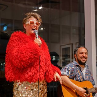 Person mit rotem, flauschigem Pullover hält ein Mikrofon, daneben sitzt eine Person mit gemustertem Hemd und spielt Gitarre