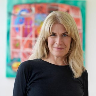 Eine lächelnde Frau mit schulterlangem, blondem Haar und schwarzem Oberteil steht vor einem farbenfrohen, unscharfen Gemälde an der Wand. Sie schaut direkt in die Kamera. Der Hintergrund ist hell und modern gestaltet.