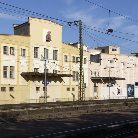 Blick auf die Aussenansicht des Franck-Areals über Gleise hinweg. Das alte Firmengebäude wird von der Sonne angestrahlt, darüber blauer Himmel.