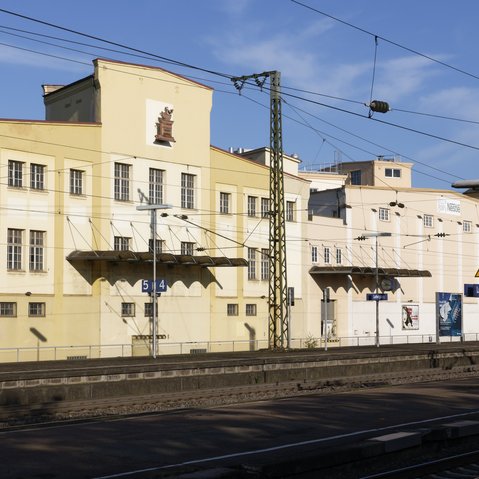 Blick auf die Aussenansicht des Franck-Areals über Gleise hinweg. Das alte Firmengebäude wird von der Sonne angestrahlt, darüber blauer Himmel.