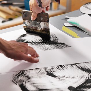 Zwei Hände arbeiten gemeinsam an einer kalligrafischen oder malerischen Komposition mit schwarzer Tinte auf weißem Papier. Ein breiter Flachpinsel wird für kräftige, dynamische Pinselstriche verwendet. Umgeben ist die Szene von Kunstmaterialien, darunter ein blauer Behälter – die Atmosphäre vermittelt konzentrierte, kreative Arbeit im Atelier.