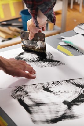 Zwei Hände arbeiten gemeinsam an einer kalligrafischen oder malerischen Komposition mit schwarzer Tinte auf weißem Papier. Ein breiter Flachpinsel wird für kräftige, dynamische Pinselstriche verwendet. Umgeben ist die Szene von Kunstmaterialien, darunter ein blauer Behälter – die Atmosphäre vermittelt konzentrierte, kreative Arbeit im Atelier.