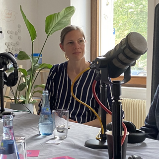 Bettina Pau, Geschäftsführerin der sitzt an einem Tisch mit Mikrofonen und Getränken, bereit für eine Podcast-Aufnahme. Sie trägt eine dunkelblaue Bluse mit weißen Streifen. Im Hintergrund sind eine grüne Zimmerpflanze und ein Fenster mit Tageslicht zu sehen.
