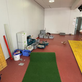 Innenraum mit rotem Boden, grünem Teppichstück und mehreren Werkzeugkisten und -behältern an der Wand