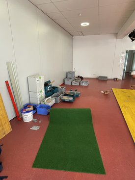 Innenraum mit rotem Boden, grünem Teppichstück und mehreren Werkzeugkisten und -behältern an der Wand