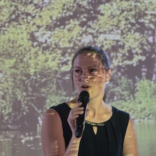 Bettina Pau, Geschäftsführerin der KulturRegion Stuttgart, während ihrer Eröffnungsrede. Sie hält ein Mikrofon in der Hand und blickt ins Publikum.