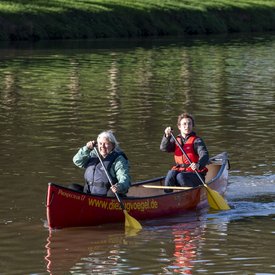 Elisabeth Maier mit männlich gelesenem Gast in einem Kanu auf einem Gewässer. Beide tragen Schwimmwesten und halten ein Paddel in den Händen.