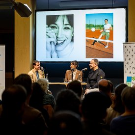 Moderator Ingmar Volkmann, Rocío Alemán und Alexander Zorniger während der Podcast Aufnahme. Alle drei sitzen auf Barhockern an Tischen und haben Mikrofone vor sich. Im Hintergrund eine Video-Leinwand, auf der Bilder gezeigt werden. Das Bild wurde über die Köpfe der Zuschauer hinweg aufgenommen.