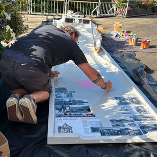 Person kniet auf dem Boden und malt mit pinker Farbe auf eine lange weiße Tafel mit schwarz-weißen historischen Fotos und Texten