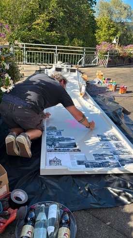 Person kniet auf dem Boden und malt mit pinker Farbe auf eine lange weiße Tafel mit schwarz-weißen historischen Fotos und Texten