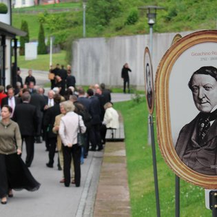 Menschenmenge auf einem Gehweg neben einer Wiese mit ovalen Porträtbildern historischer Persönlichkeiten auf Ständern