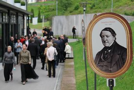 Menschenmenge auf einem Gehweg neben einer Wiese mit ovalen Porträtbildern historischer Persönlichkeiten auf Ständern