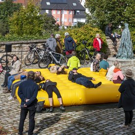 Einige Teilnehmer*innen der Bustour liegen oder sitzen auf einem großen, gelben Kissen im Schlosshof. Die Personen scheinen die Sonne zu genießen, sich zu unterhalten oder der Hörstation zu lauschen.