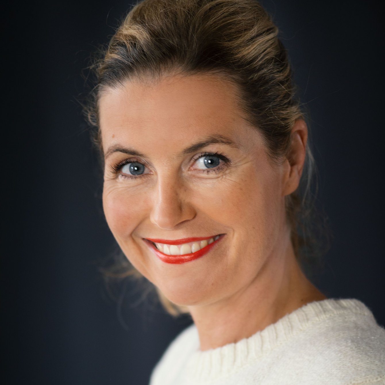 Portrait von Nicola Brakamp. Nicola Brakamp lächelt in die Kamera. Ihre Haare sind hochgesteckt, sie trägt einen weißen Wollpulli und auffallend roten Lippenstift.