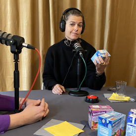 Gäst*in Sara Dahme während der Aufnahme des Podcast. Sie sitzt an einem Tisch, trägt Kopfhörer und hat ein Tischmikrofon vor sich stehen. In der Hand hält sie einen typischen Schuldrink, den sie skeptisch betrachtet.