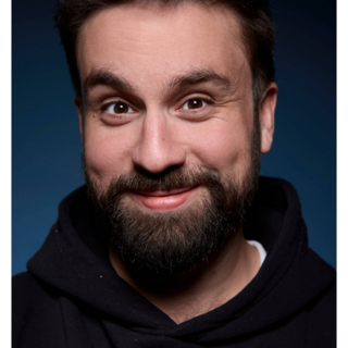 ein junger Mann mit schwarzen Haaren und Bart lächelt verschmitzt in die Kamera