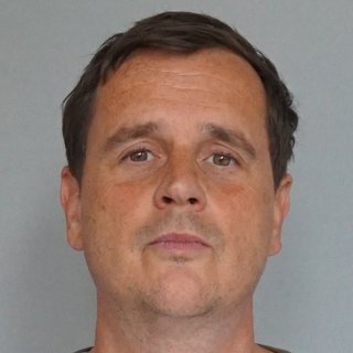 Ein Mann mit kurzen, dunklen Haaren blickt ruhig in die Kamera. Er trägt ein weißes T-Shirt unter einem olivgrünen Pullover oder einer Strickjacke. Der Hintergrund ist neutral grau.