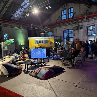 Besucher sitzen und stehen in einem industriellen Raum, um eine Gaming-Präsentation bei einem Kunst-im-Game-Symposium zu verfolgen.