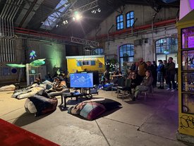 Besucher sitzen und stehen in einem industriellen Raum, um eine Gaming-Präsentation bei einem Kunst-im-Game-Symposium zu verfolgen.