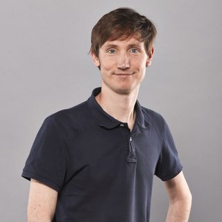 Ein junger Mann mit kurzen, braunen Haaren steht vor einem neutral grauen Hintergrund. Er trägt ein dunkelblaues Poloshirt, schaut freundlich in die Kamera und hat eine Hand in die Hüfte gestützt