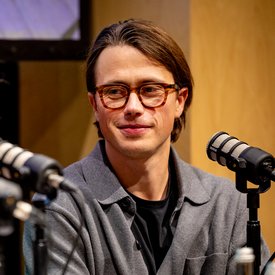 Nils Unterharnscheidt (Agentur Societas) hat braunes, schulterlanges Haar und trägt einen grauen Pullover über schwarzem T-Shirt. Er sitzt vor Mikrofonen und lächelt.