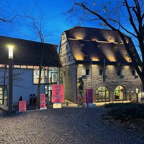 Abendaufnahme eines historischen Gebäudes in Böblingen mit beleuchtetem Plakat, das zur Ausstellung ‚Schlamassel Tov‘ einlädt. Verschiedene pinkfarbene Plakate sind vor dem Gebäude sichtbar.