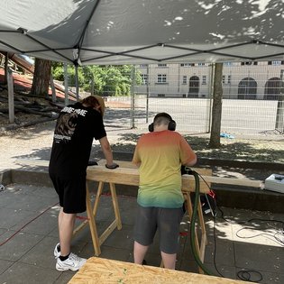 Zwei Personen unter einem Pavillon, eine steht und arbeitet an einem Holzbrett, die andere trägt Gehörschutz und hält ein Elektrowerkzeug