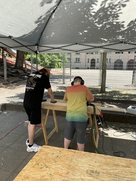 Zwei Personen unter einem Pavillon, eine steht und arbeitet an einem Holzbrett, die andere trägt Gehörschutz und hält ein Elektrowerkzeug