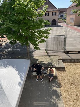 Drei Personen sitzen auf einer niedrigen Mauer neben einem Baum und einem weißen Zelt, im Hintergrund ein umzäunter Platz mit Gebäuden