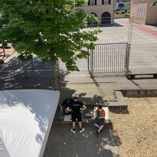 Drei Personen sitzen auf einer niedrigen Mauer neben einem Baum und einem weißen Zelt, im Hintergrund ein umzäunter Platz mit Gebäuden