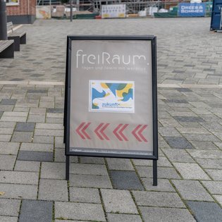 Auf einem Gehweg steht ein Hinweisschild mit der Aufschrift ‚freiRaum – tagen und feiern mit Weitblick‘. Darunter befindet sich ein farbenfrohes Plakat mit abstrakten Formen und dem Text ‚zukunftkultur – Die Kulturinitiative der KulturRegion Stuttgart‘. Drei rote Pfeile zeigen nach rechts. Im Hintergrund sind Sitzbänke und Gebäude zu sehen.