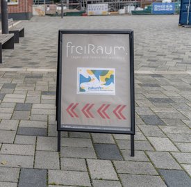 Auf einem Gehweg steht ein Hinweisschild mit der Aufschrift ‚freiRaum – tagen und feiern mit Weitblick‘. Darunter befindet sich ein farbenfrohes Plakat mit abstrakten Formen und dem Text ‚zukunftkultur – Die Kulturinitiative der KulturRegion Stuttgart‘. Drei rote Pfeile zeigen nach rechts. Im Hintergrund sind Sitzbänke und Gebäude zu sehen.