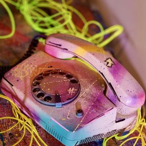 Altes Wählscheibentelefon mit bunter Sprühfarbe und gelben Kabeln auf bemaltem Untergrund.