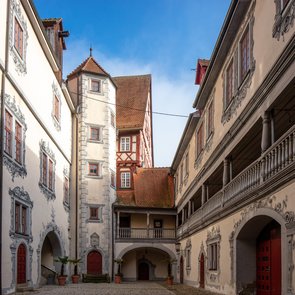 Innenhof eines historischen Schlosses mit weiß verputzten Fassaden, roten Türen und Fenstern sowie einem blauen Himmel im Hintergrund
