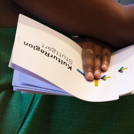 Hand mit dunkler Hautfarbe blättert in einem Buch mit dem Titel 'Kulturregion Stuttgart' auf grünem Stoffhintergrund