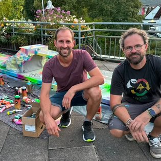 Zwei Männer sitzen auf dem Boden neben einem bunten, selbst gestalteten Minigolfhindernis mit Spraydosen und Kartons um sie herum