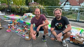 Zwei Männer sitzen auf dem Boden neben einem bunten, selbst gestalteten Minigolfhindernis mit Spraydosen und Kartons um sie herum