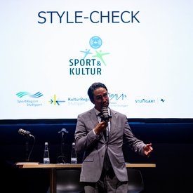 Dr. Torben Giese im grauen Anzug hält Mikrofon vor einer Leinwand mit dem Text 'STYLE-CHECK' und Logos von SportRegion Stuttgart, KulturRegion Stuttgart, StadtPalais, Stuttgart.