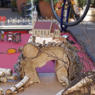 Miniaturkirche auf einem Holzstamm mit Birkenholzscheiben und einem Fahrrad im Hintergrund