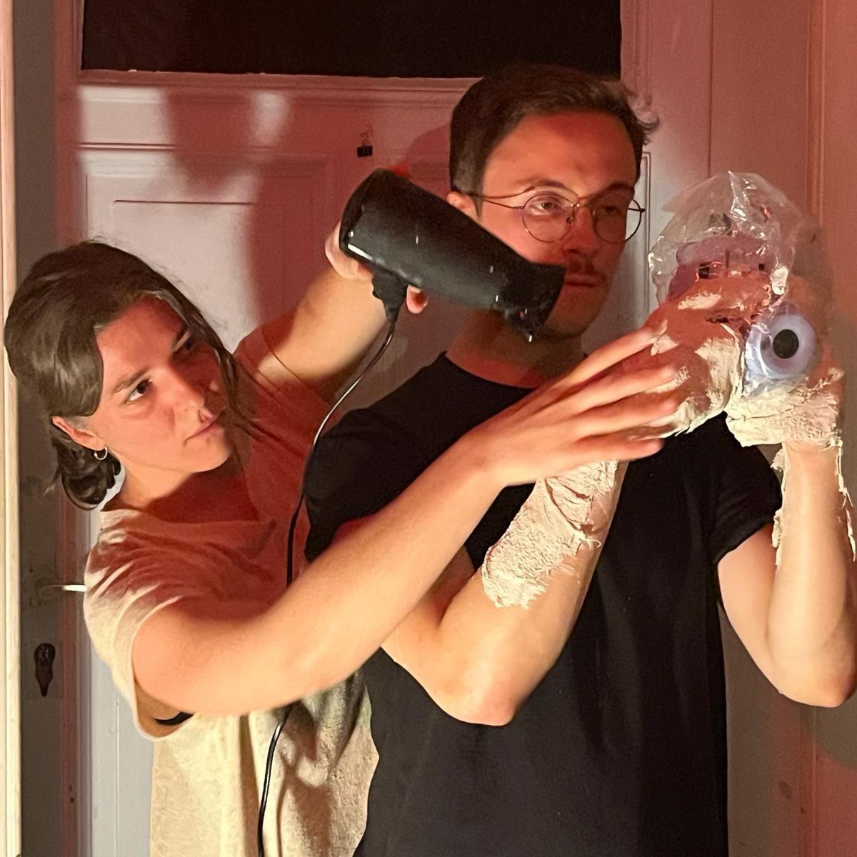 Selbstportrait von Emilia Dorr und Tyler Cunningham. Emilia Dorr hält einen Fön in der Hand, den sie auf etwas richtet, was Tyler Cunningham in der Hand hält. 