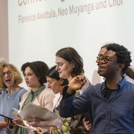Neo Muyanga und der Projektchor beim Singen. Neo Muyanga gibt mit der rechten Hand Zeichen für den Einsatz der Sänger.