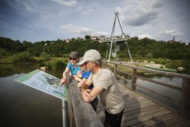 Eine Familie, gekleidet wie Wanderer, schaut sich eine Infotafel auf einem Holzsteg an. Um den Holzsteg herum sieht man eine Auenlandschaft.