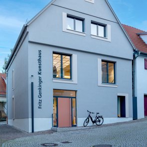 Moderne Außenansicht des Fritz Genkinger Kunsthauses in Marbach. Das hellgraue Gebäude mit Satteldach hat drei Stockwerke und schlichte, rechteckige Fenster. An der Fassade steht in vertikaler Schrift »Fritz Genkinger Kunsthaus«. Vor dem Eingang steht ein schwarzes Fahrrad, der Vorplatz ist mit Kopfsteinpflaster bedeckt. Der Himmel ist klar und blau.